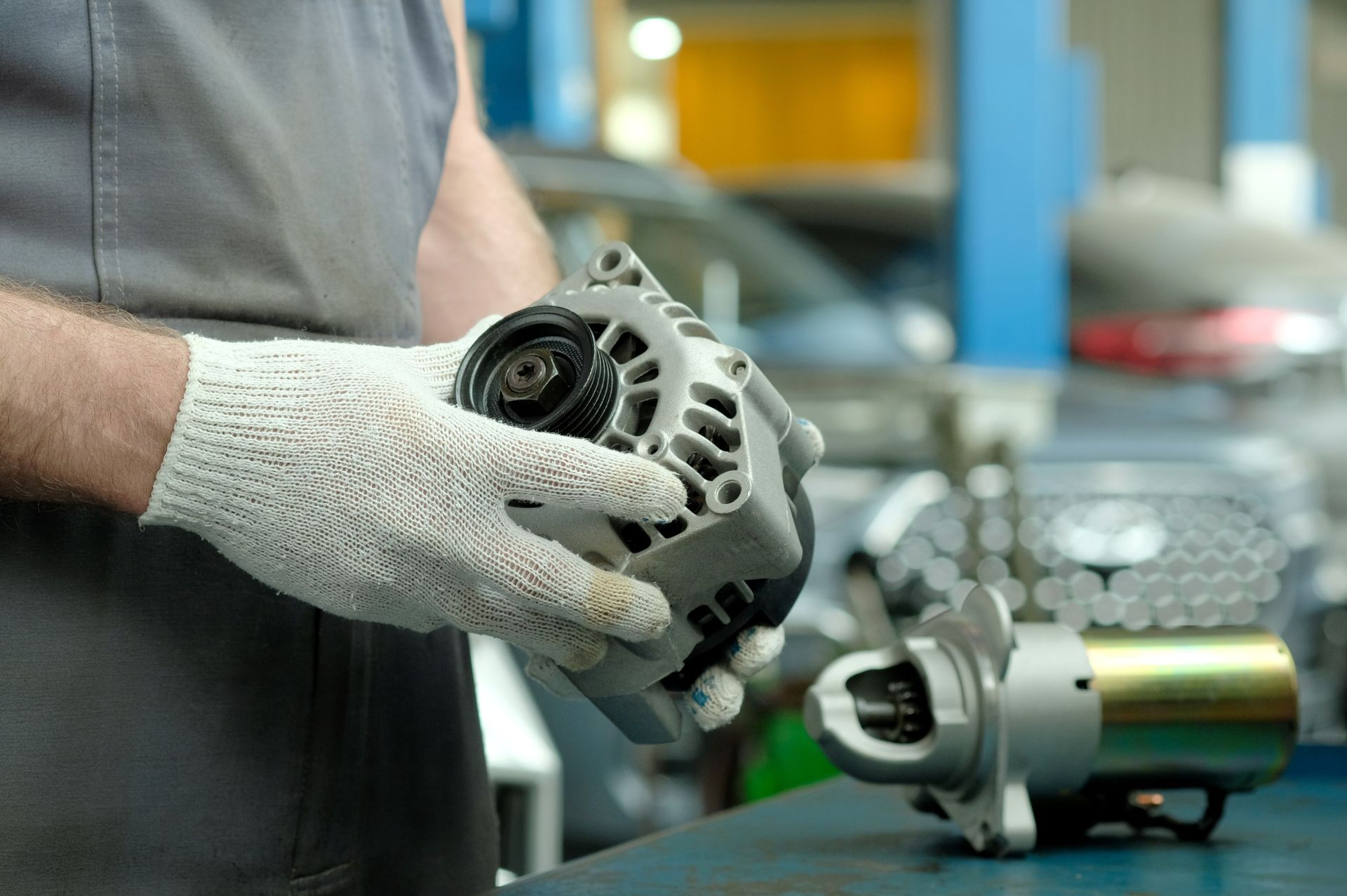 Gloved hands holding a car alternator in a workshop, with another alternator on the table.