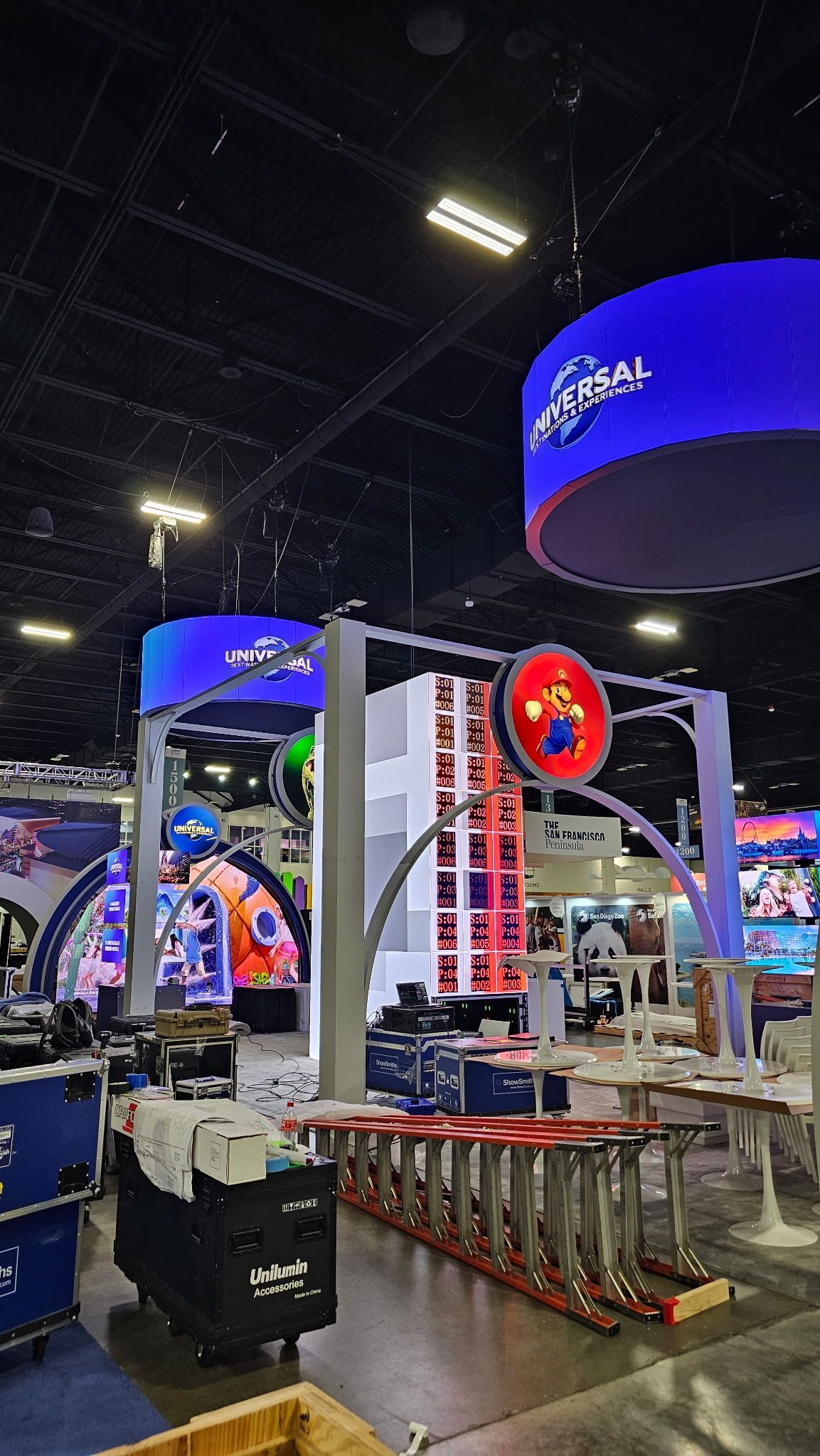 Trade show booth with large blue hanging screens, product displays, and equipment in an indoor exhibition hall being assembled