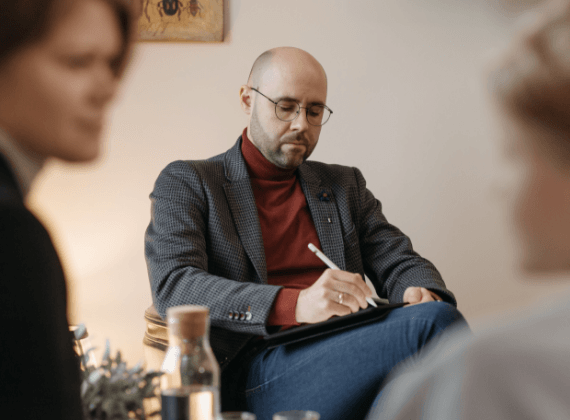 Een man in pak zit voor een groep mensen en schrijft op een klembord.
