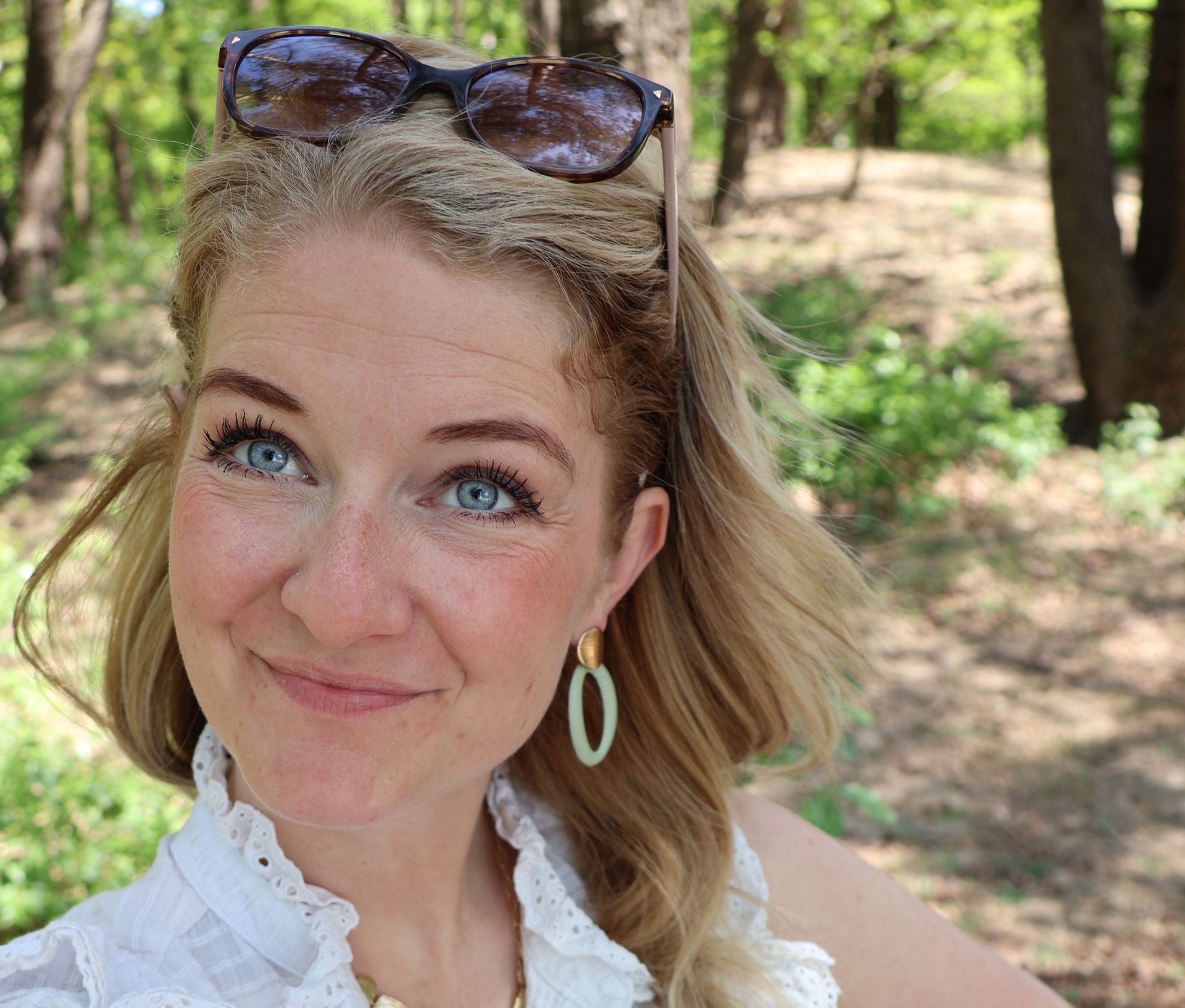 Blonde vrouw met een zonnebril op haar hoofd, lichtblauwe ogen, glimlachend in een bosrijke omgeving, oorbellen dragend.