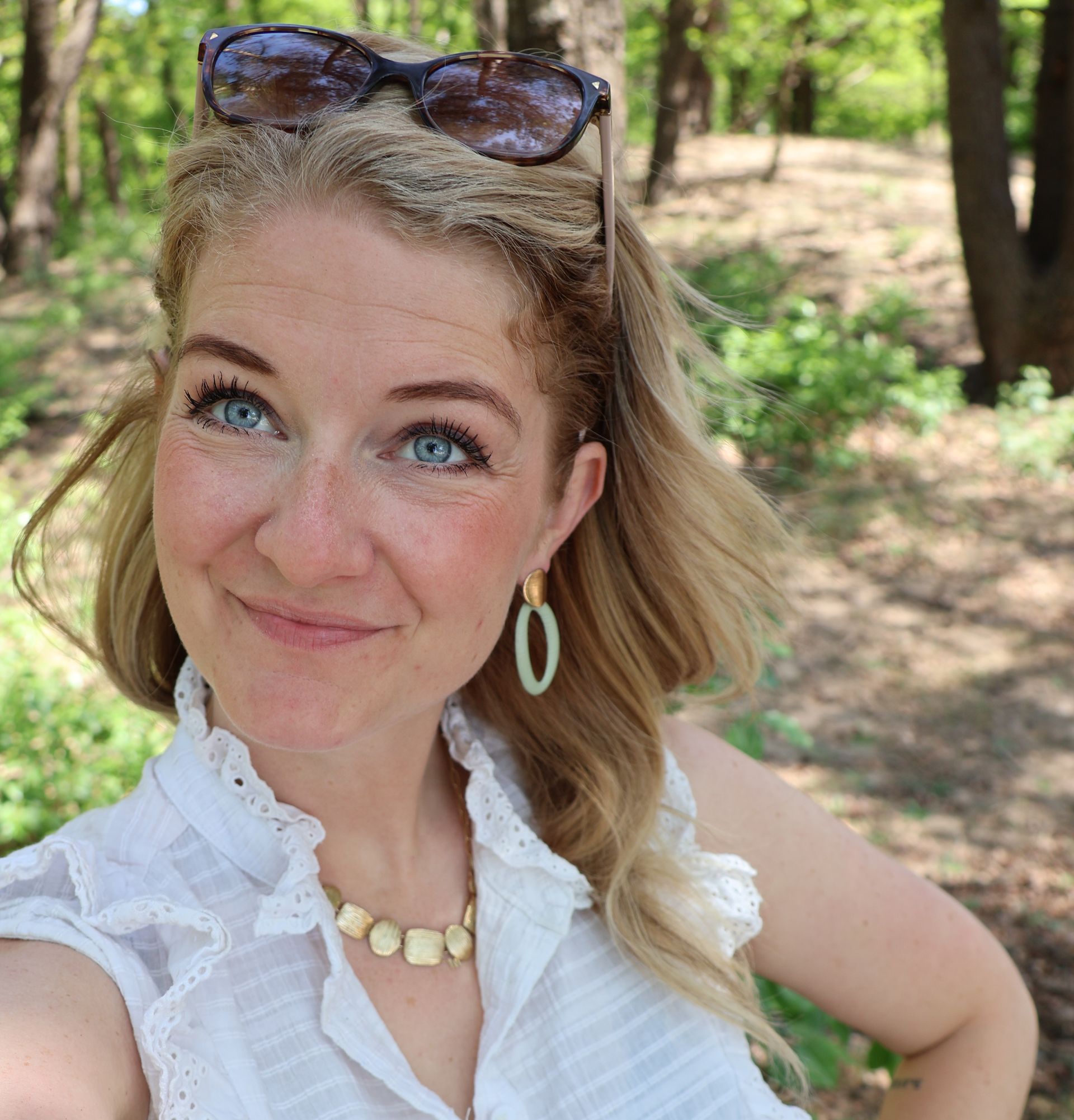 Een vrouw met blauwe ogen lacht naar de camera in een bosrijke omgeving. Ze draagt ​​een zonnebril. Ze draagt ​​een wit overhemd en een ketting.