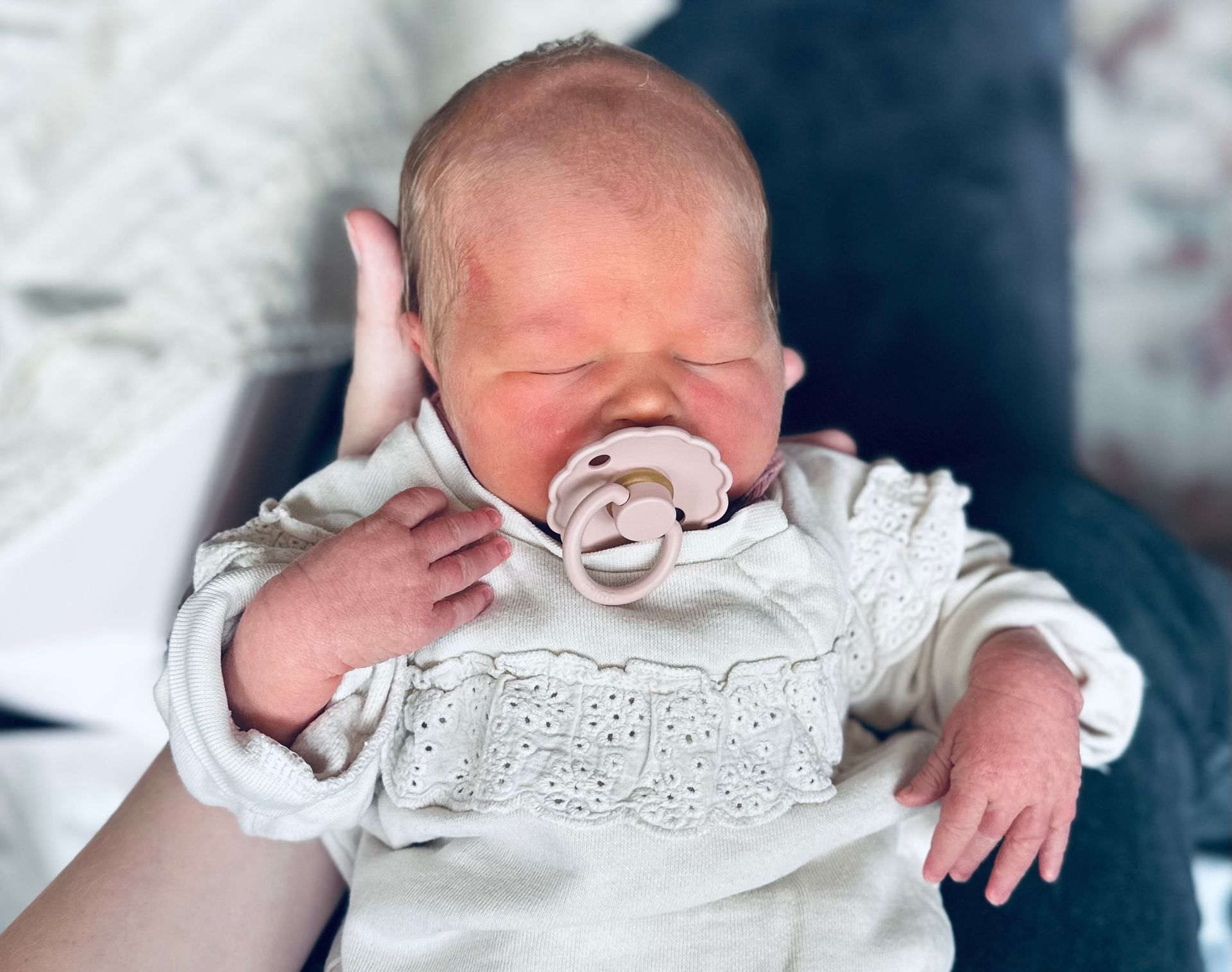 Pasgeboren baby met een fopspeen, vastgehouden in iemands armen, gekleed in een witte trui.