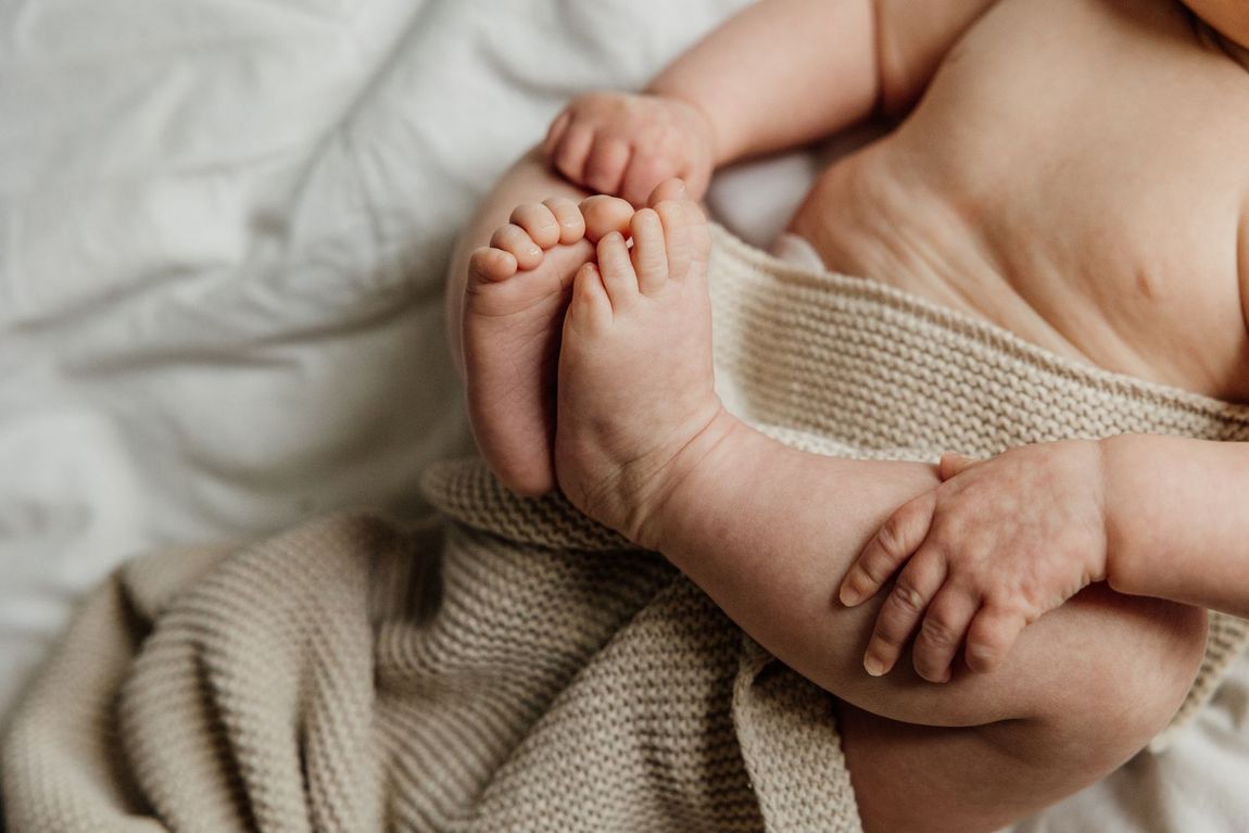 Baby ligt op bed met een beige deken, benen en voeten zichtbaar.