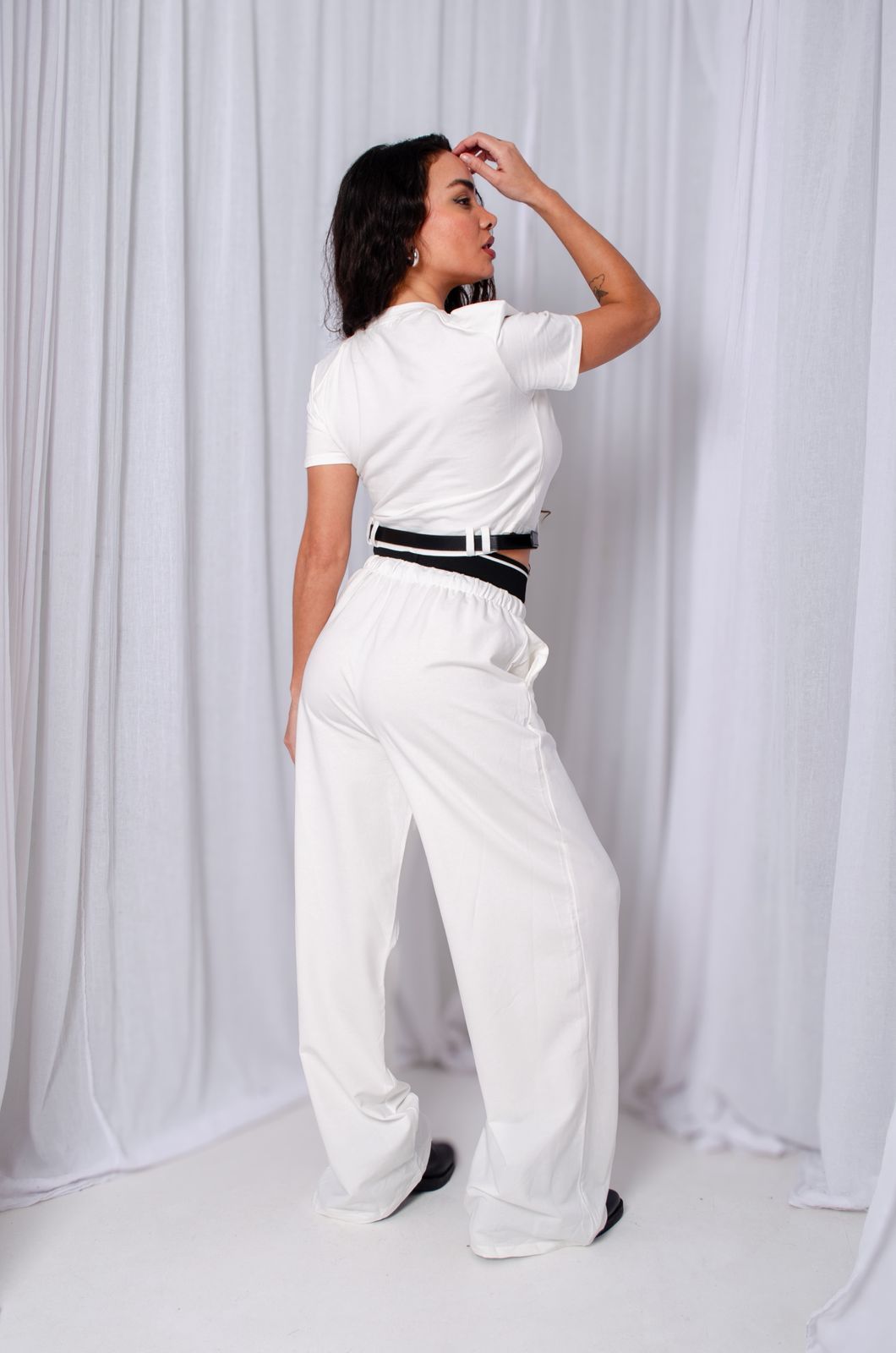 Mujer vestida de blanco con cinturón negro, de pie frente a unas cortinas blancas.