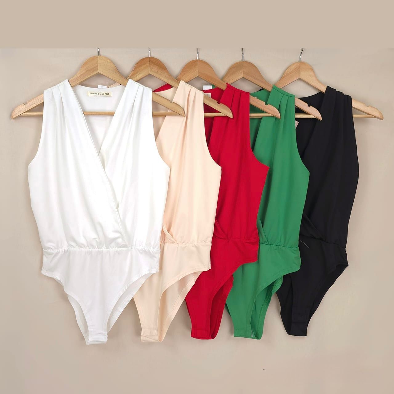 Cinco bodies sin mangas en blanco, melocotón, rojo, verde y negro, colgados en perchas de madera.