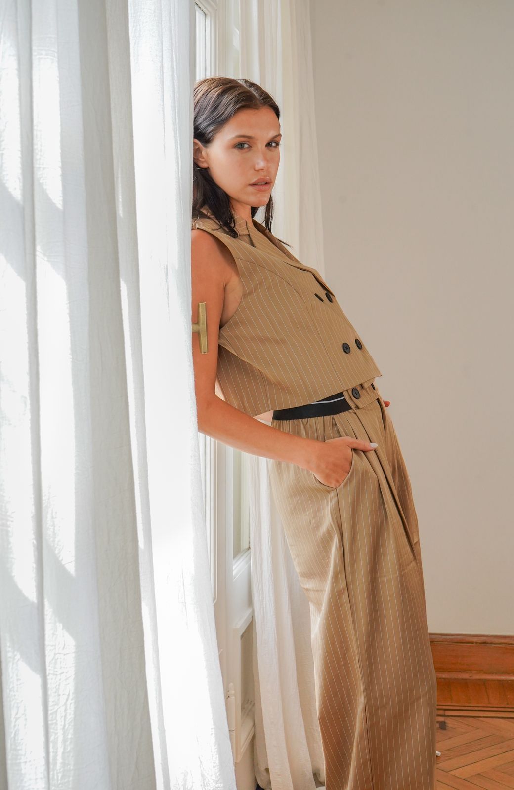 Mujer con traje de rayas color canela se apoya en una cortina blanca, mirando a la cámara.