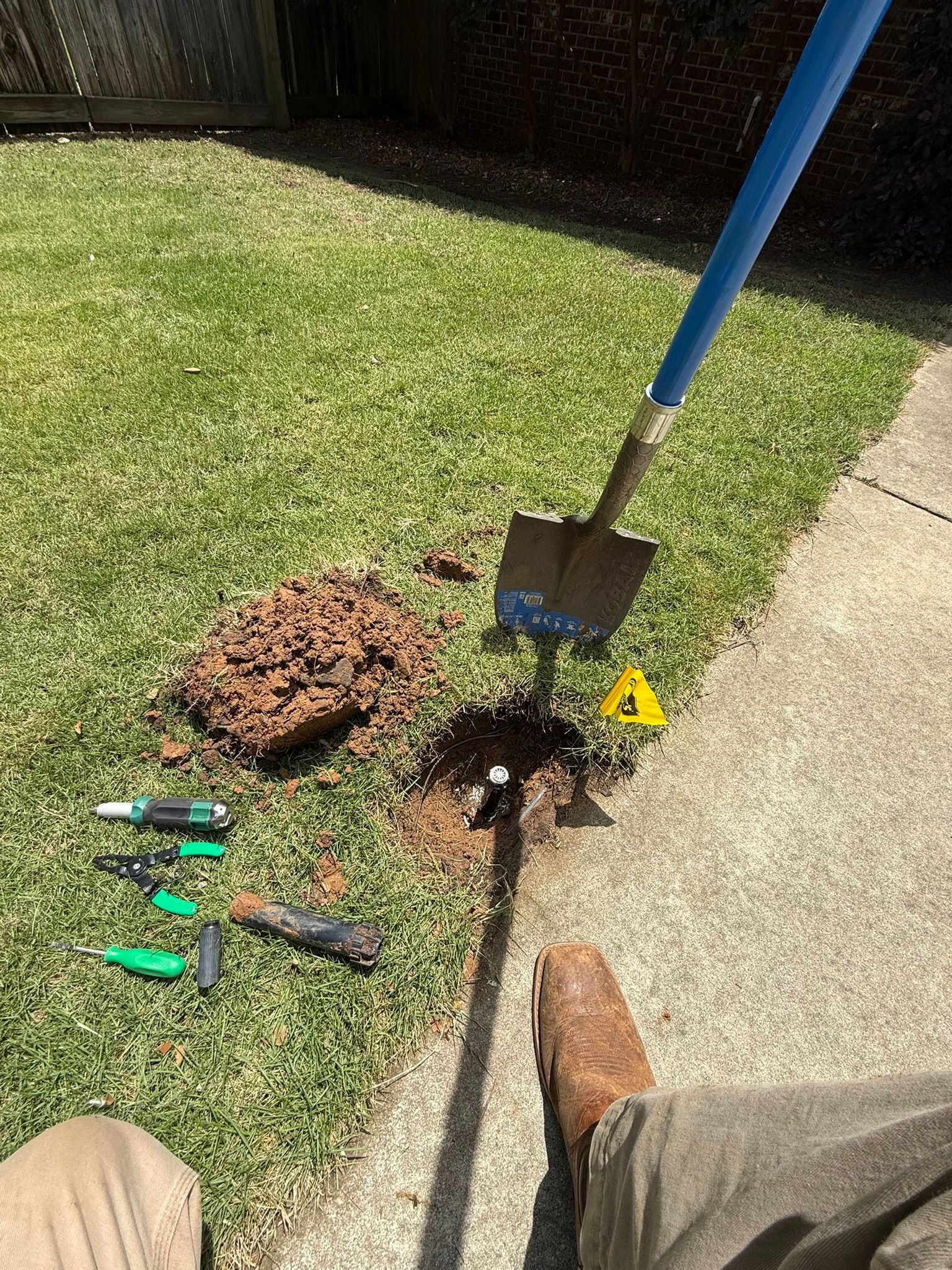 A shovel and disturbed soil with tools on grass near a sidewalk.