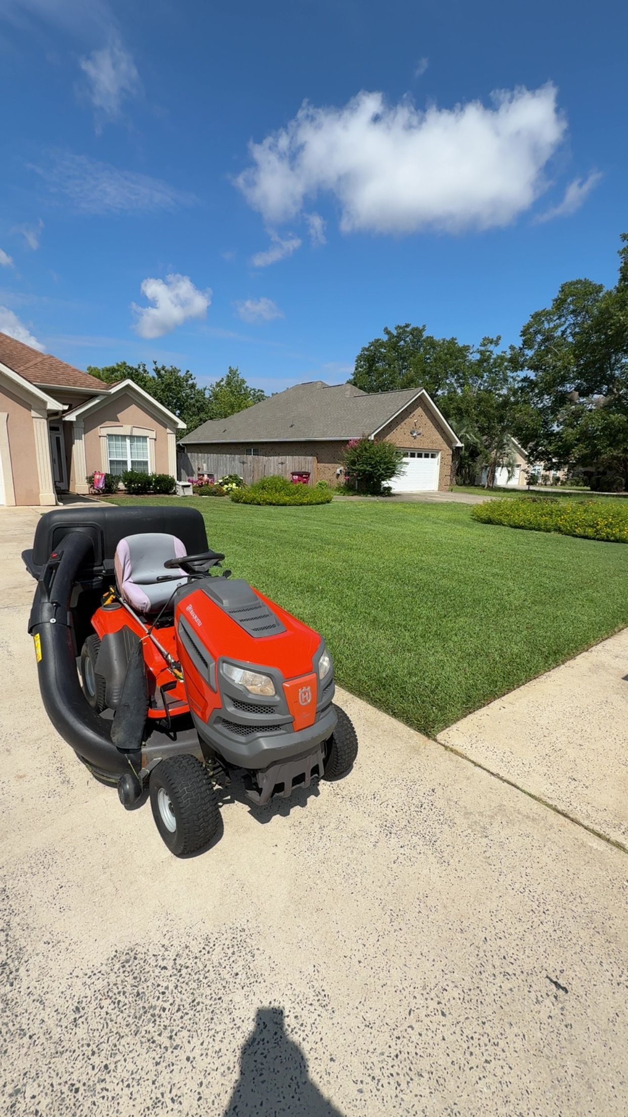 Riding lawnmower with collection bin on a paved driveway, next to a freshly cut lawn and houses, under a blue sky.