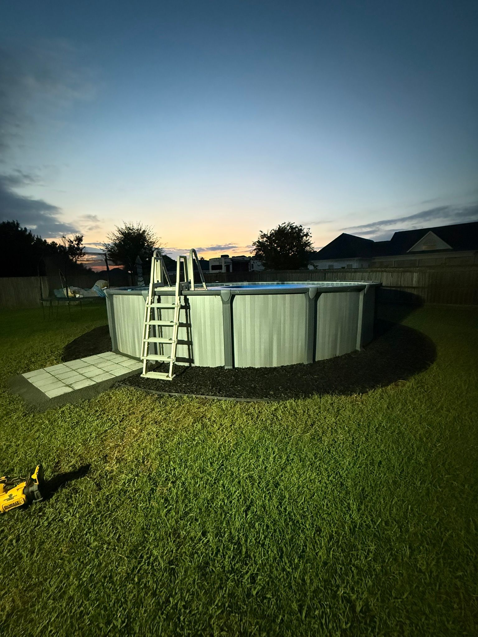 Above-ground pool in a grassy yard with a ladder, surrounded by black mulch and a gray walkway at dusk.