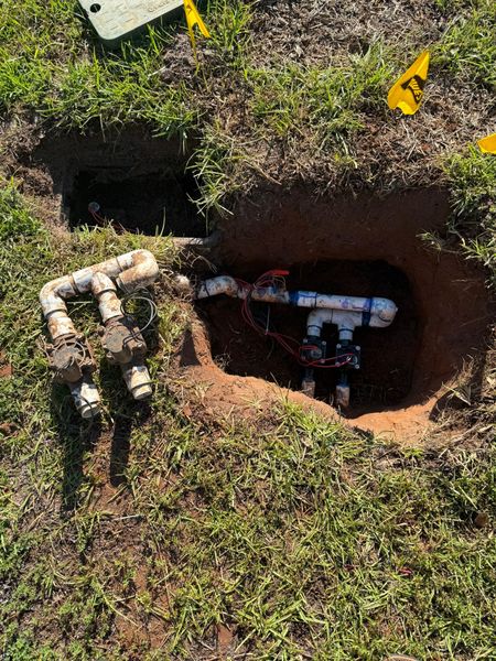 Open ground revealing PVC pipes and sprinkler valves in a grassy yard. Yellow flags mark locations.