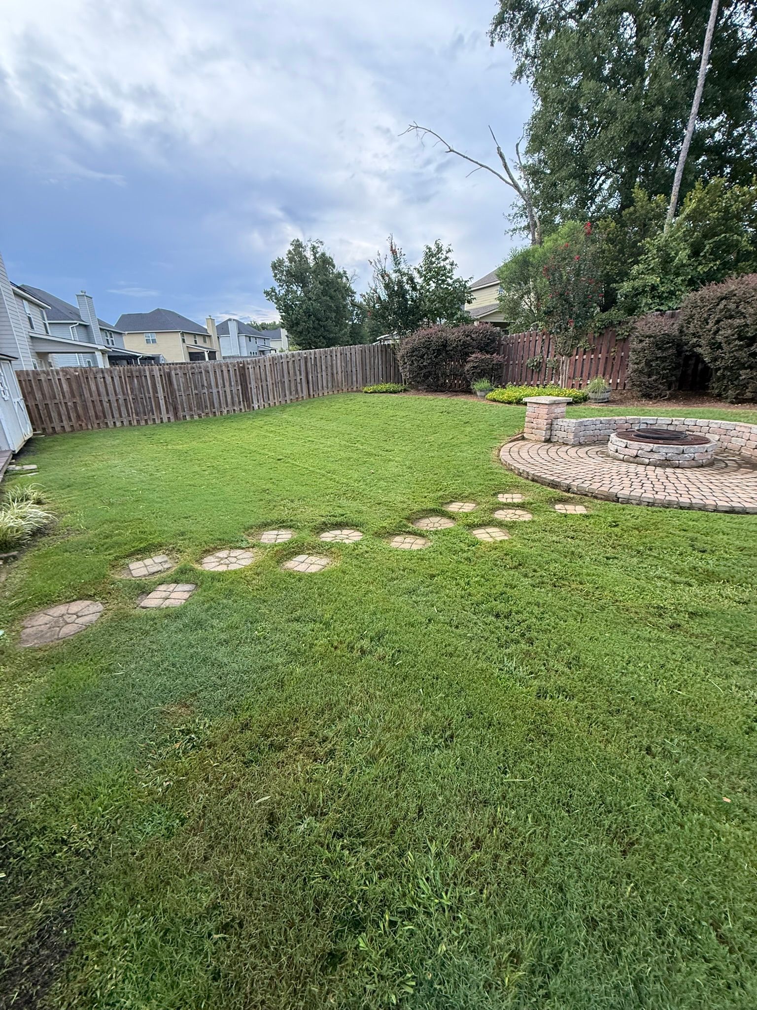 A grassy backyard with a stone path, fence, and fire pit under a cloudy sky.