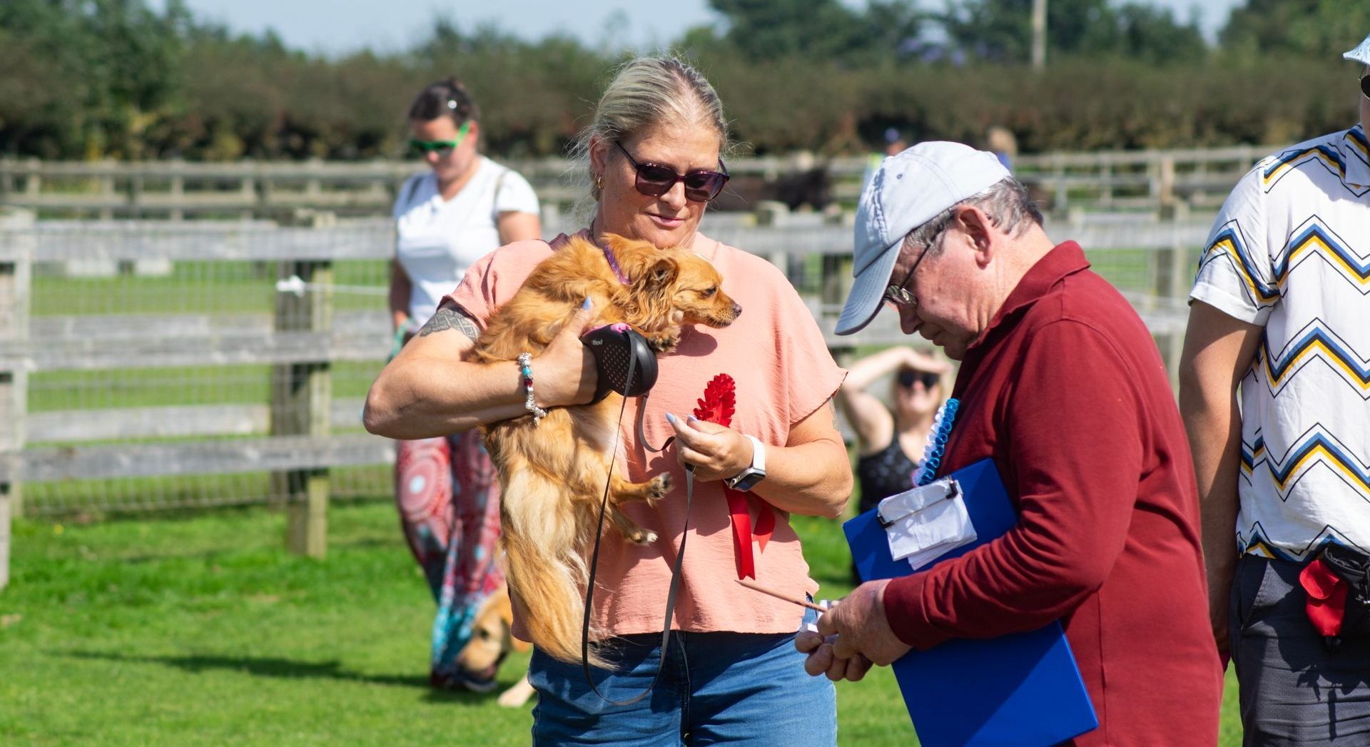 A woman holding a small, brown dog, and a man looking at a clipboard, outdoors at an event.