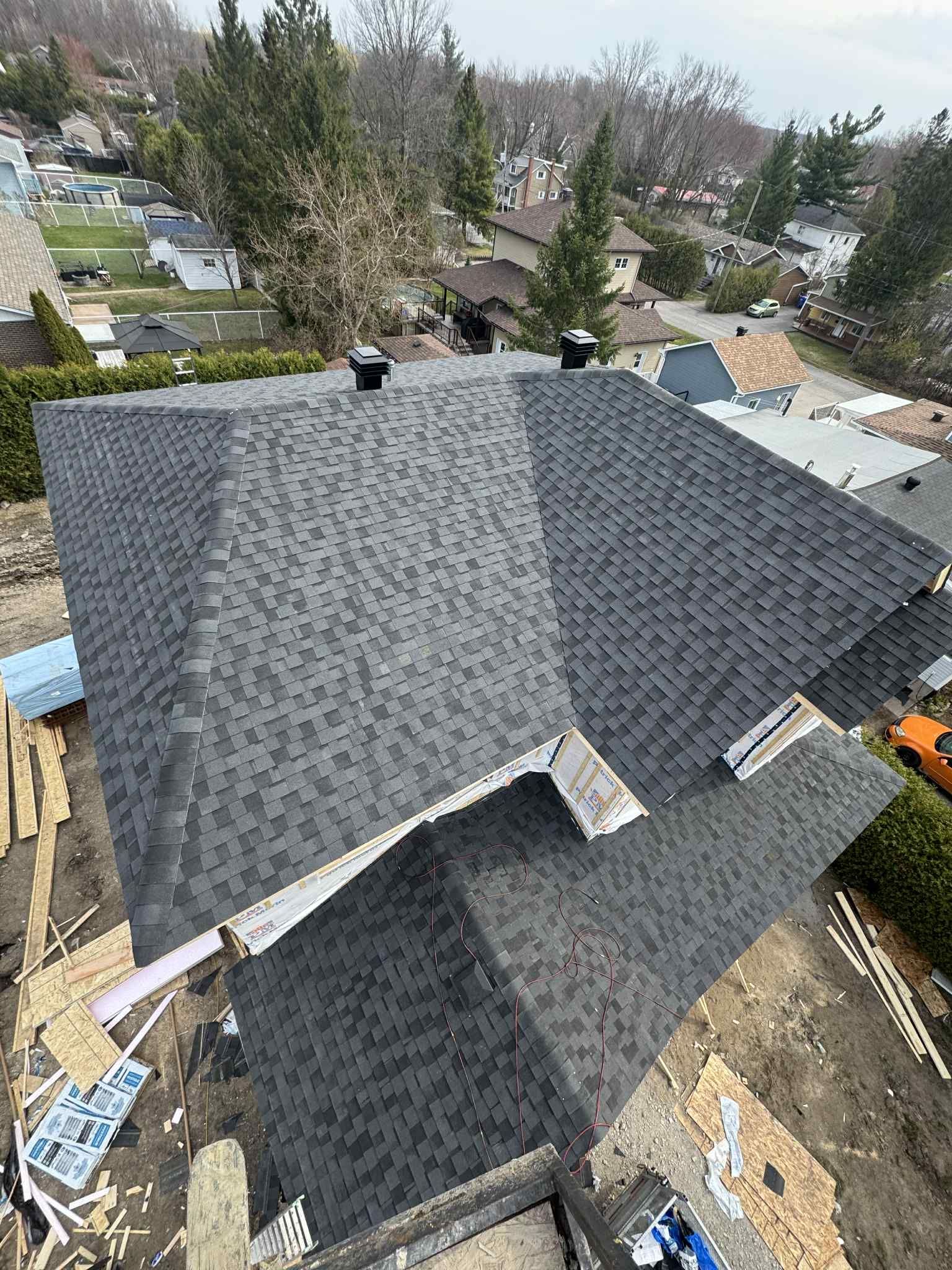 Vue aérienne du toit d'une maison avec des bardeaux gris foncé, deux aérations noires et des arbres environnants.