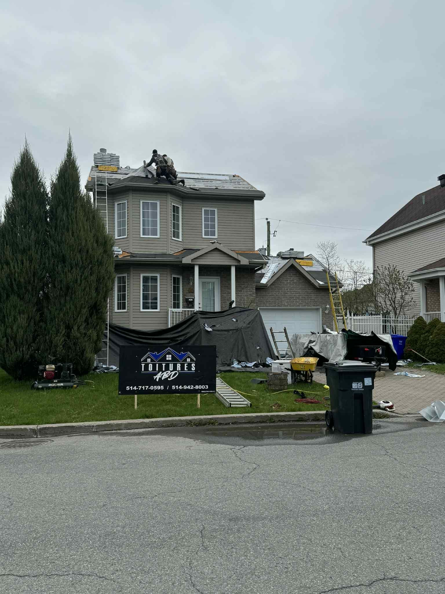 Maison en cours de réfection de toiture ; ouvriers sur le toit, bâche sur la pelouse, banderole devant.