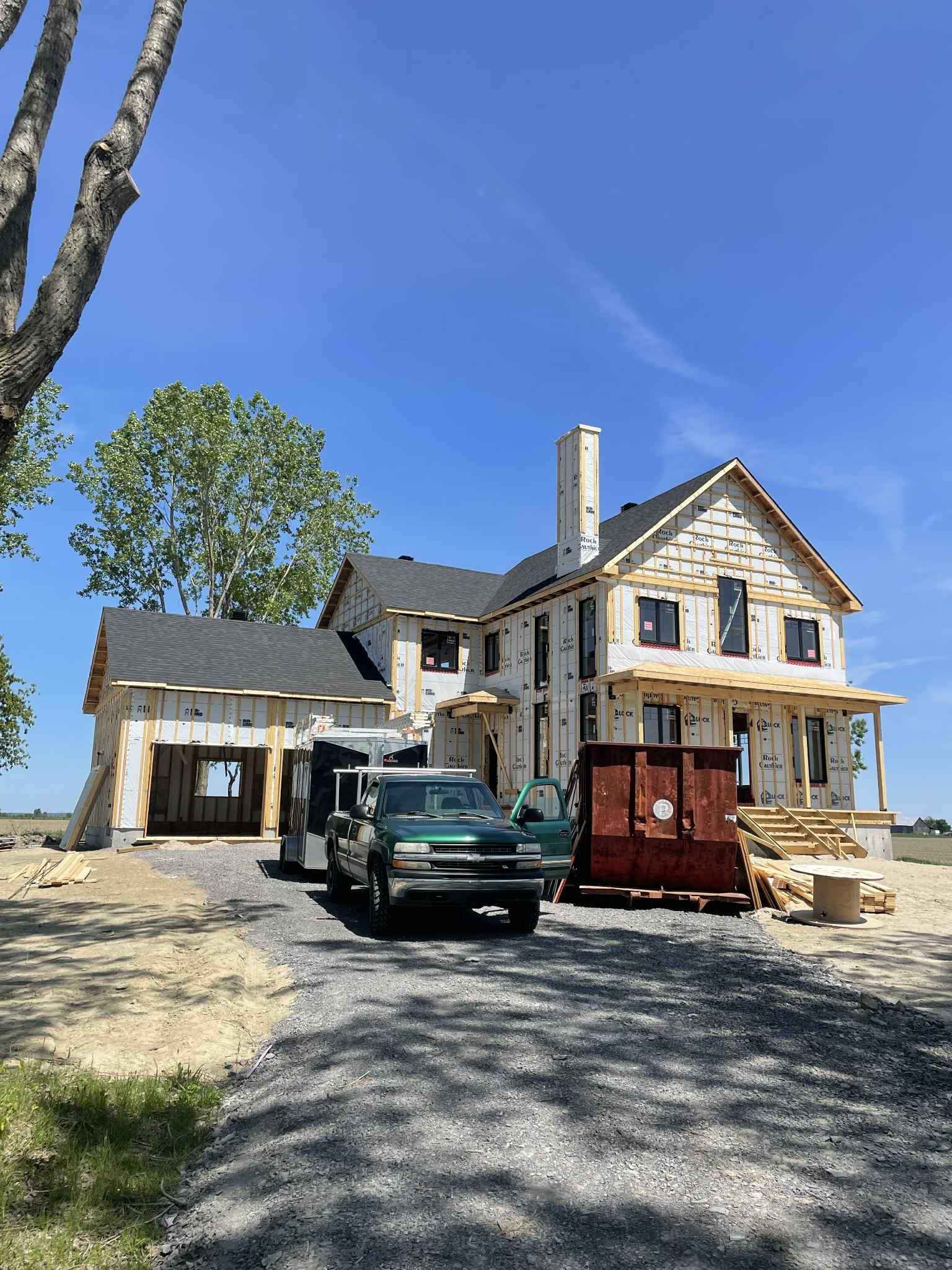 Maison en construction, à ossature bois, toiture noire, et camions de chantier sur une allée de gravier sous un ciel bleu.