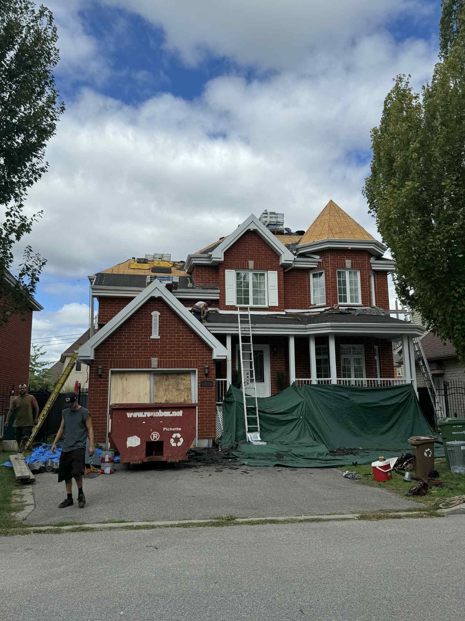 Maison en construction ; façade en briques rouges, toiture partiellement démontée. Ouvriers présents, ciel nuageux.