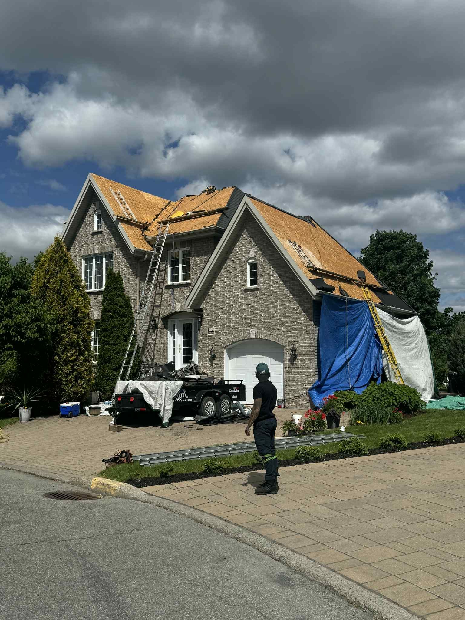 Maison au toit partiellement exposé, recouvert de bâches. Un ouvrier se tient sur l'allée.