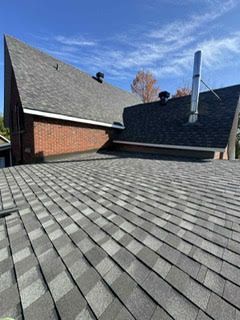 Toit en bardeaux gris foncé avec cheminée et mur de briques sur fond de ciel bleu.
