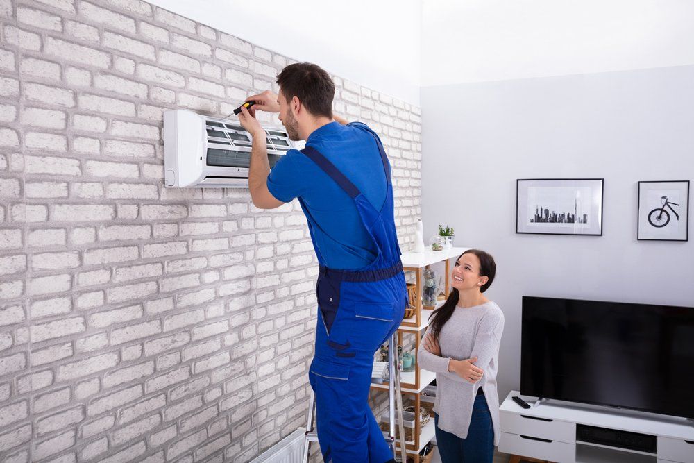 Woman Looking at Male Technician Repairing Air Conditioner — South Bend, IN — Ernsperger Air Conditioning & Heating Inc