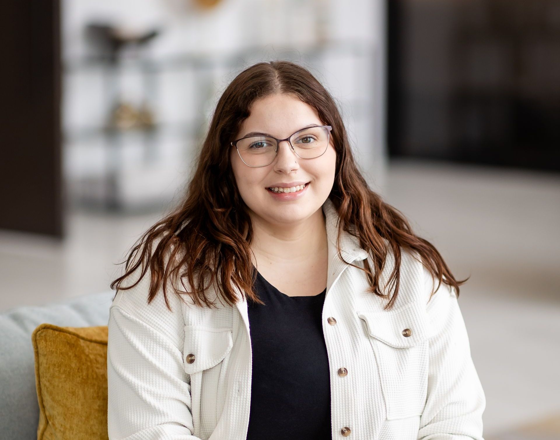 A young woman wearing glasses and a white jacket is sitting on a couch.