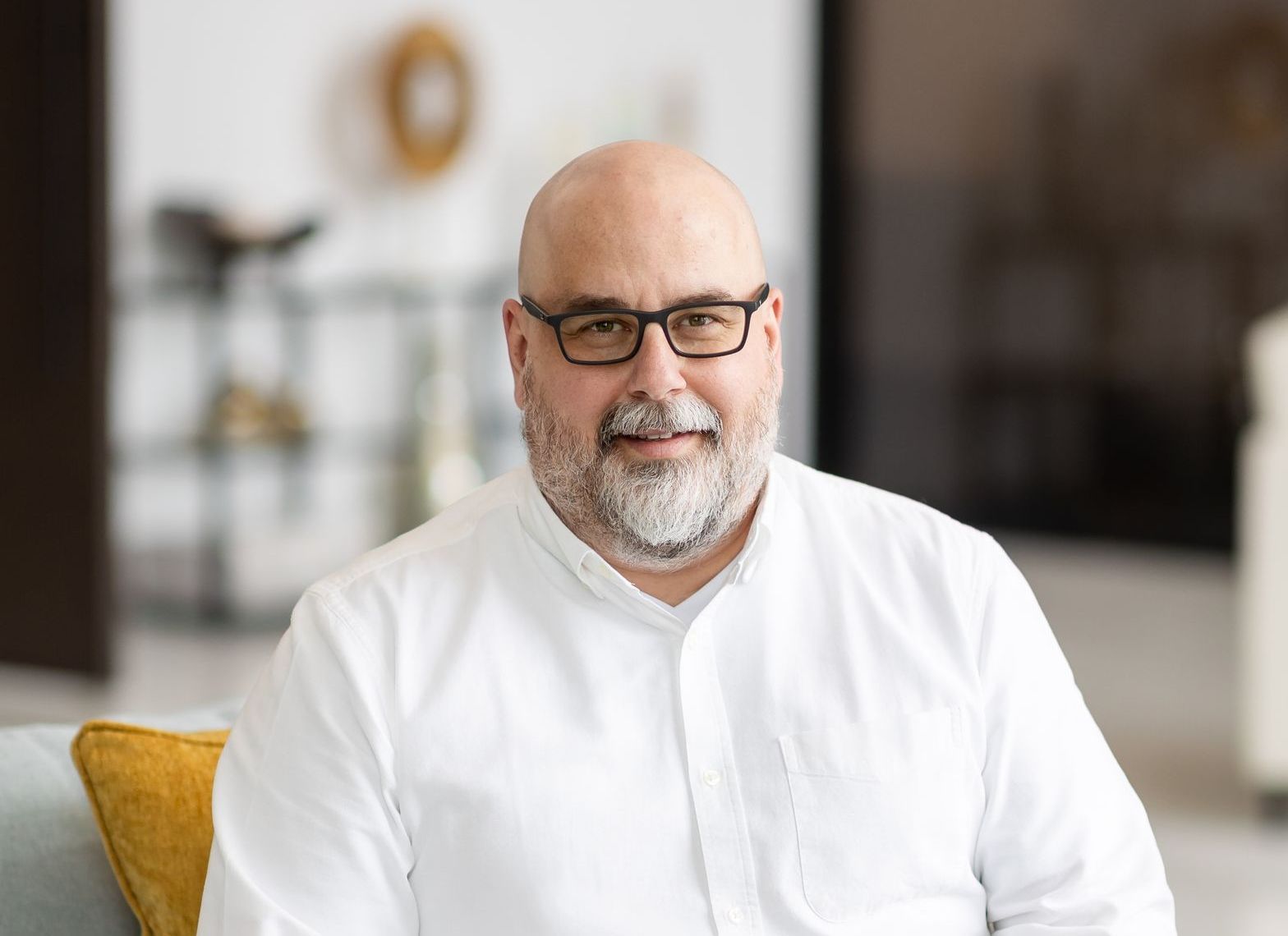 A bald man with a beard and glasses is sitting on a couch.