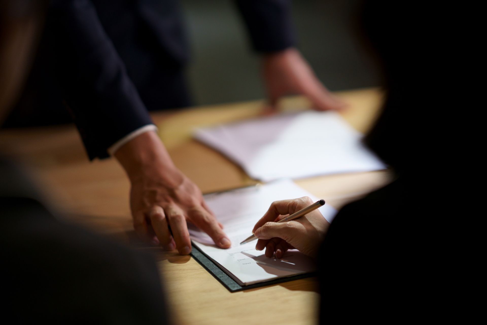 Person signs legal document as another hand points, in a professional office setting.