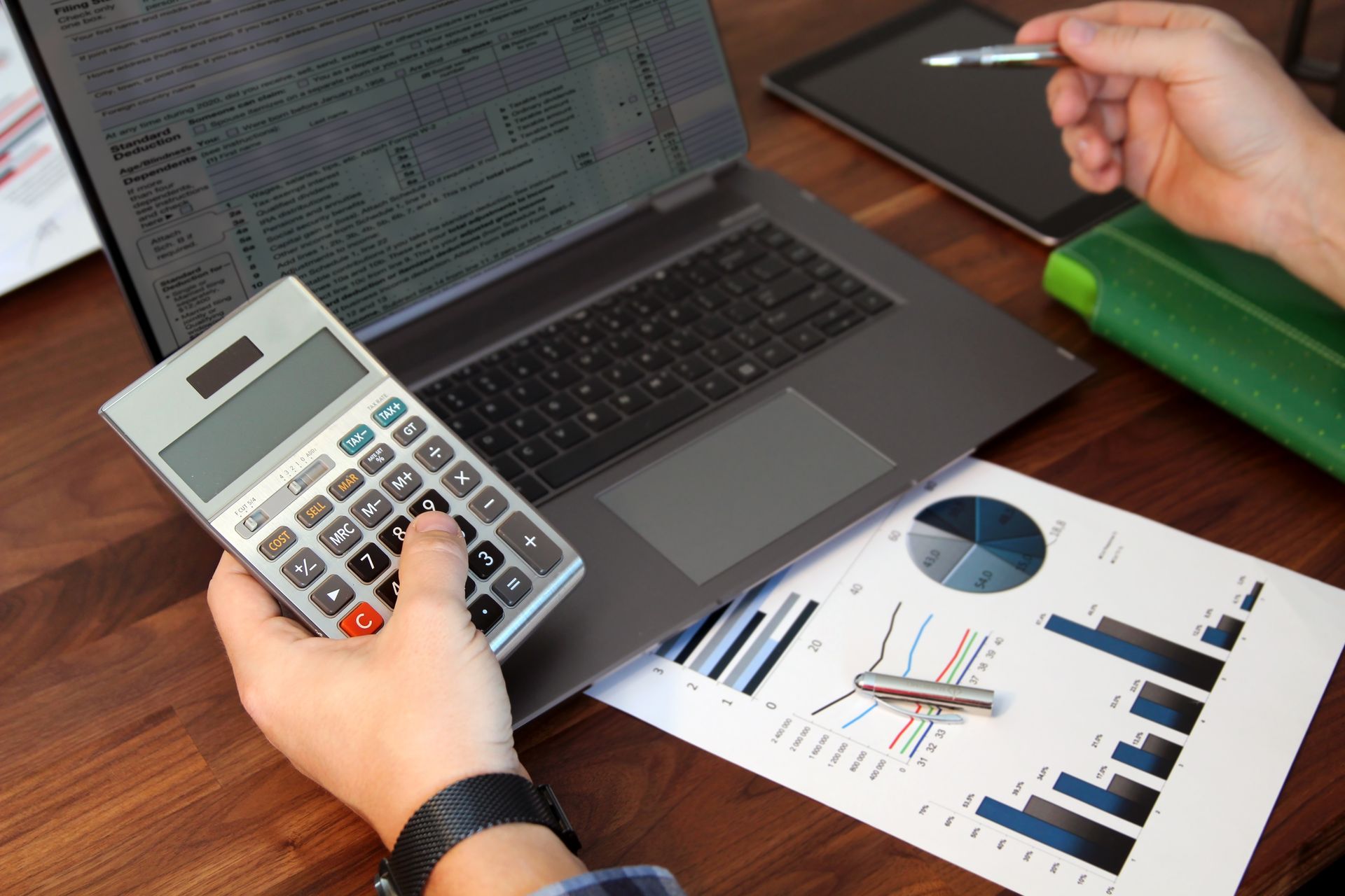 Person calculating numbers with a calculator at a desk with a laptop and financial reports.