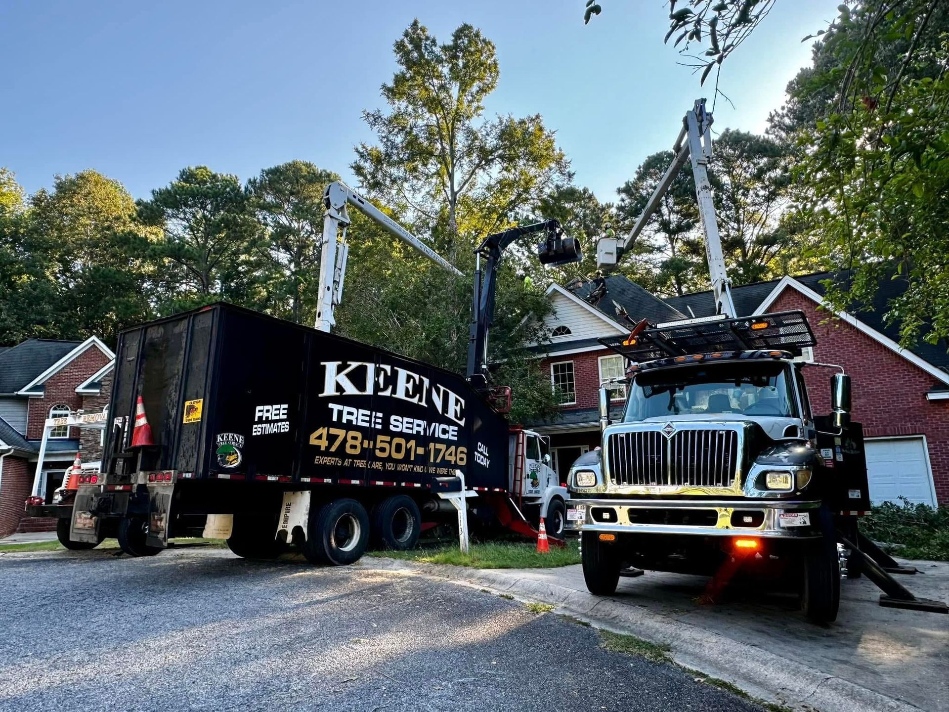Trucks from Keene Tree Service near a house, with one truck's lift arm extended toward a tree.