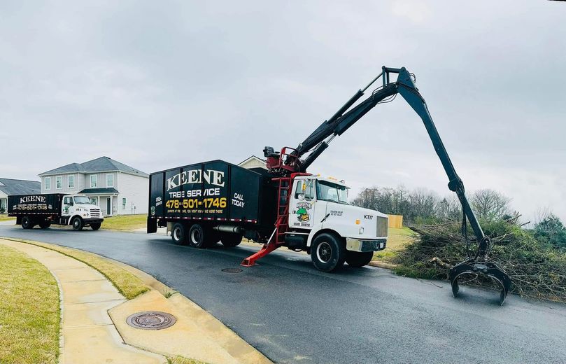 A crane truck loads debris from a roadside into a container. Another truck follows on the street.