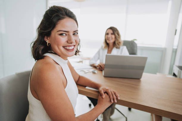 A woman is sitting at a table with a laptop and smiling.