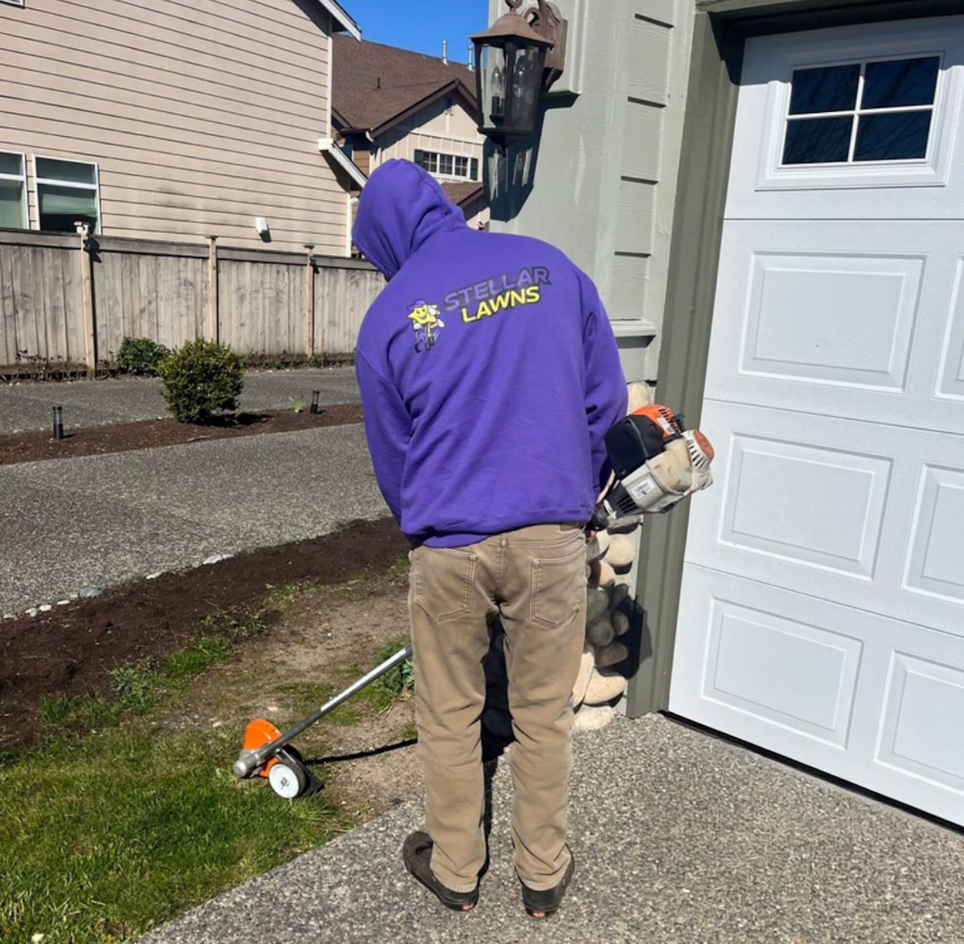 Person in purple hoodie using a trimmer beside a garage and driveway.