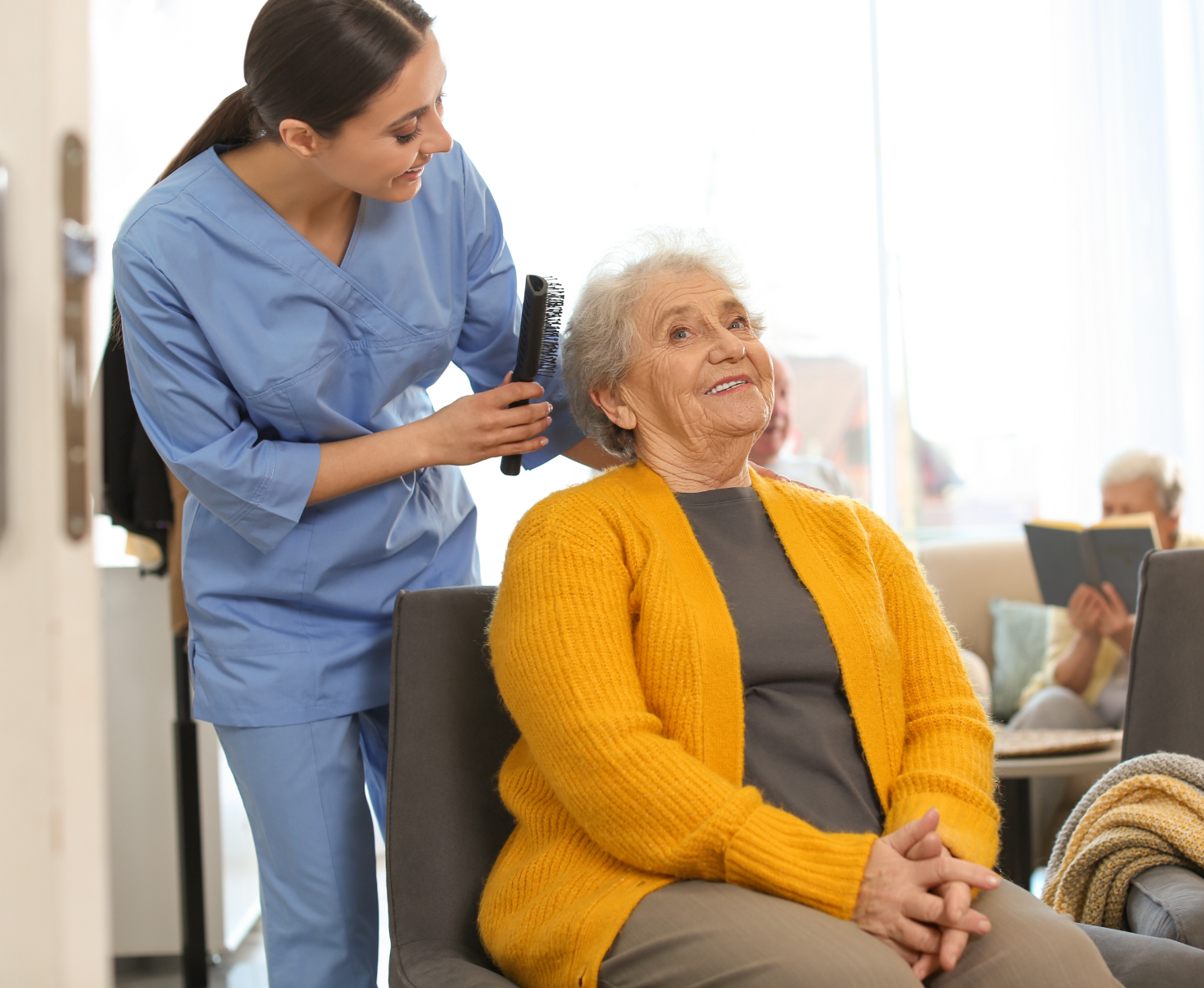 A caregiver brushes an elderly woman's hair. The woman smiles while seated in a lounge chair.