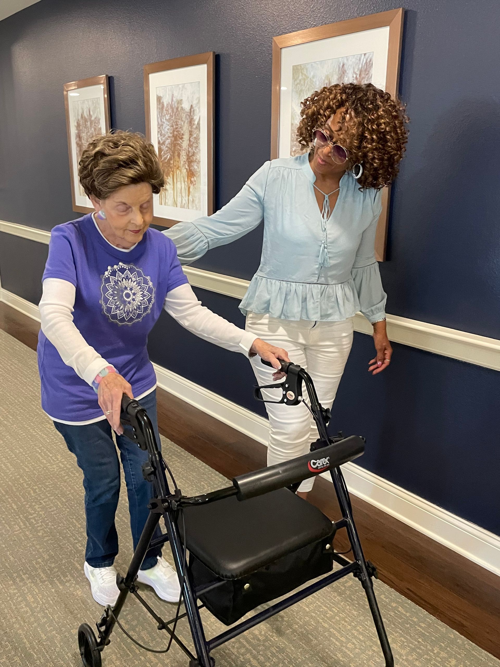 A woman is helping another woman with a walker in a hallway.