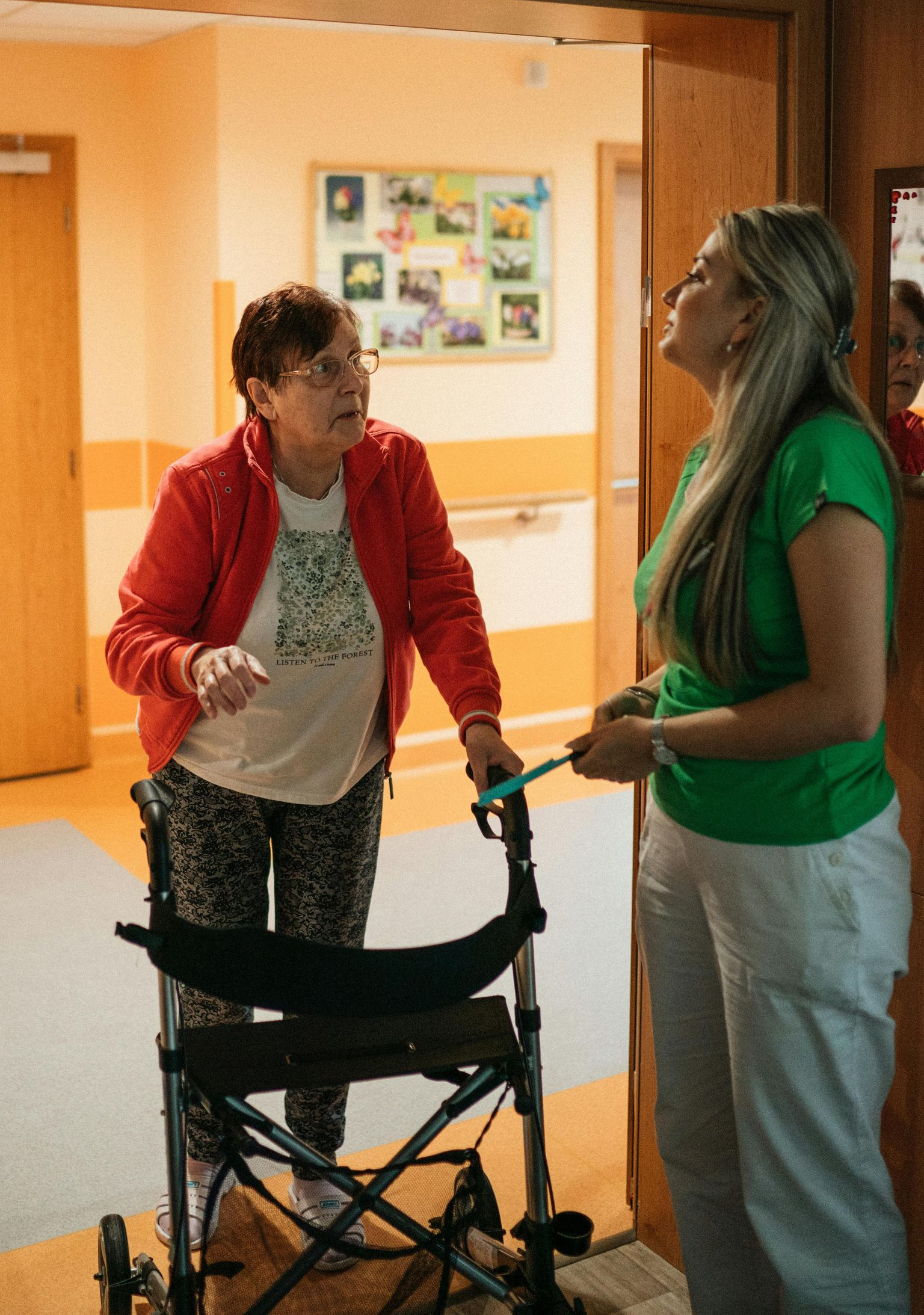 An elderly woman using a walker talks to a younger woman in a hallway. Both are smiling.