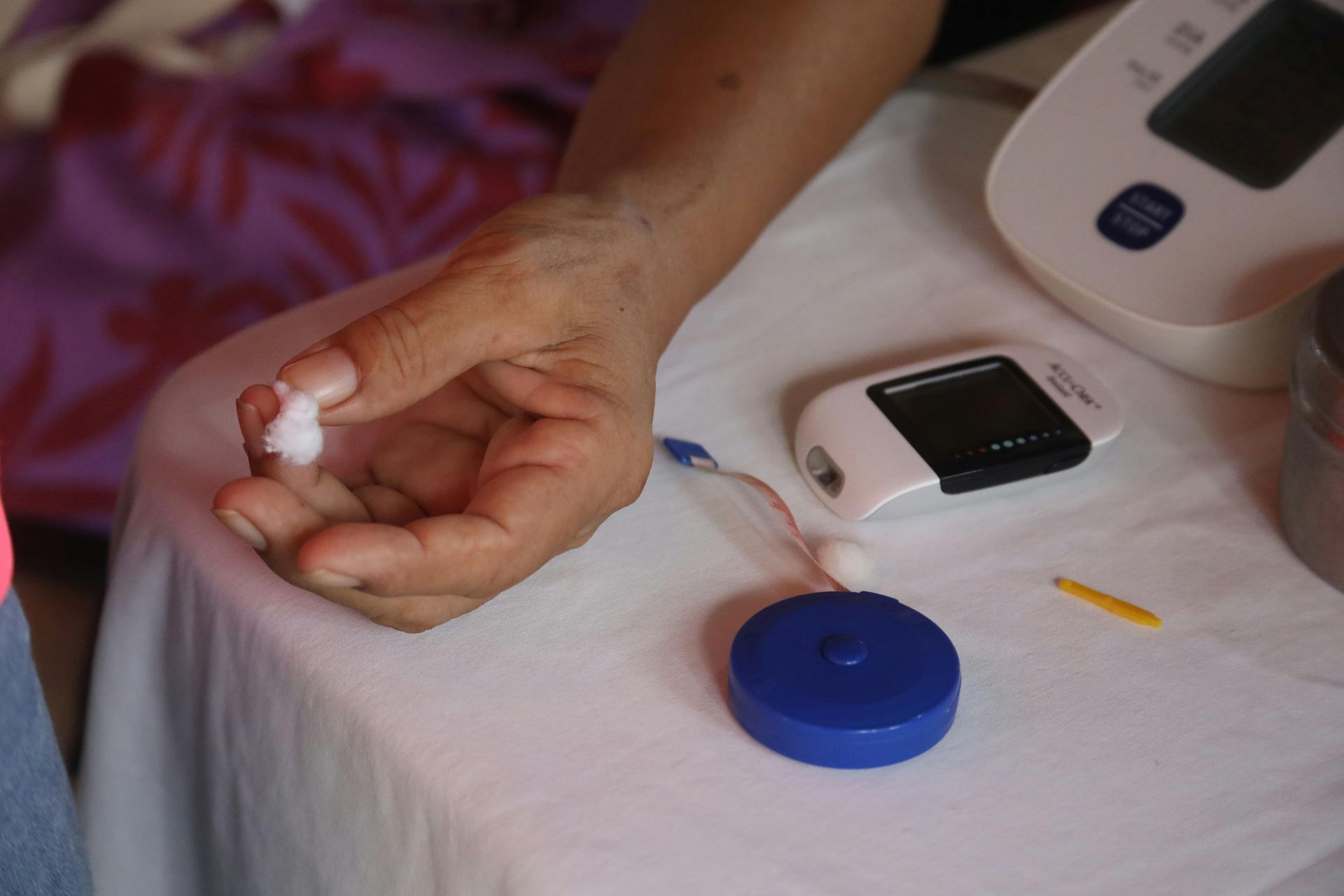 Person's hand holding cotton swab, likely after a blood test, resting on a table with a blood pressure monitor and other medical supplies.