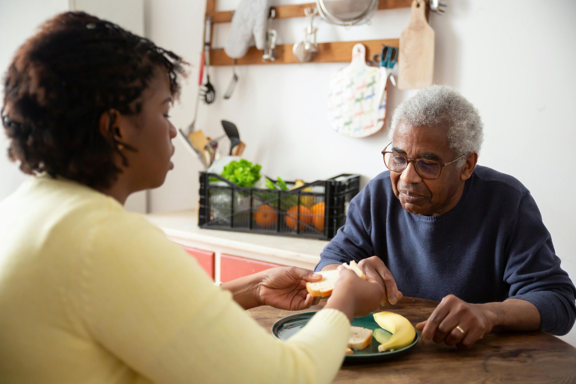 A Black woman and an elderly Black man at a table. She holds a sandwich slice for him, he looks down, hands clasped.