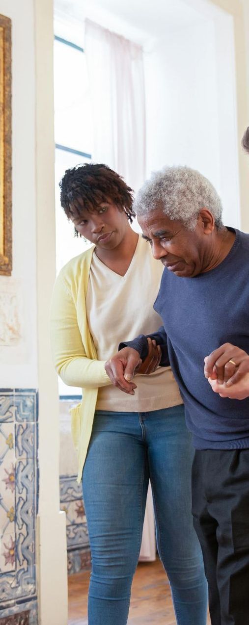 Woman assisting an elderly man walking indoors. She gently guides him, both looking focused and attentive.