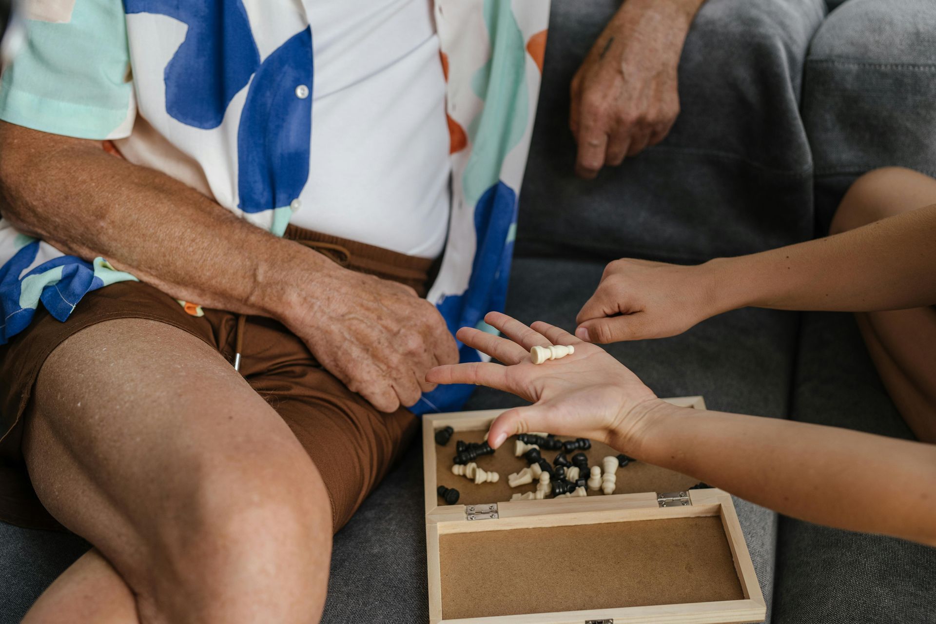 An elderly person and a younger person playing chess on a sofa. The younger person holds chess pieces in their hand.