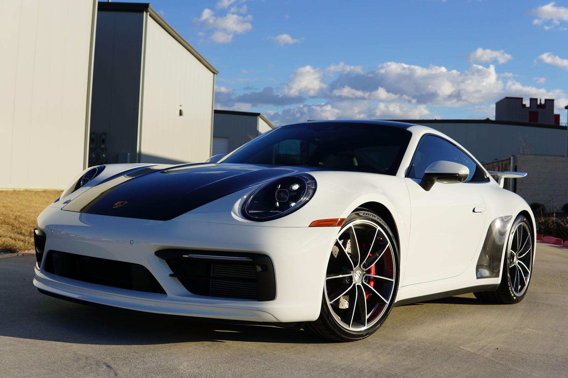White Porsche 911 coupe with black hood and black wheels, parked in front of a building on a sunny day.