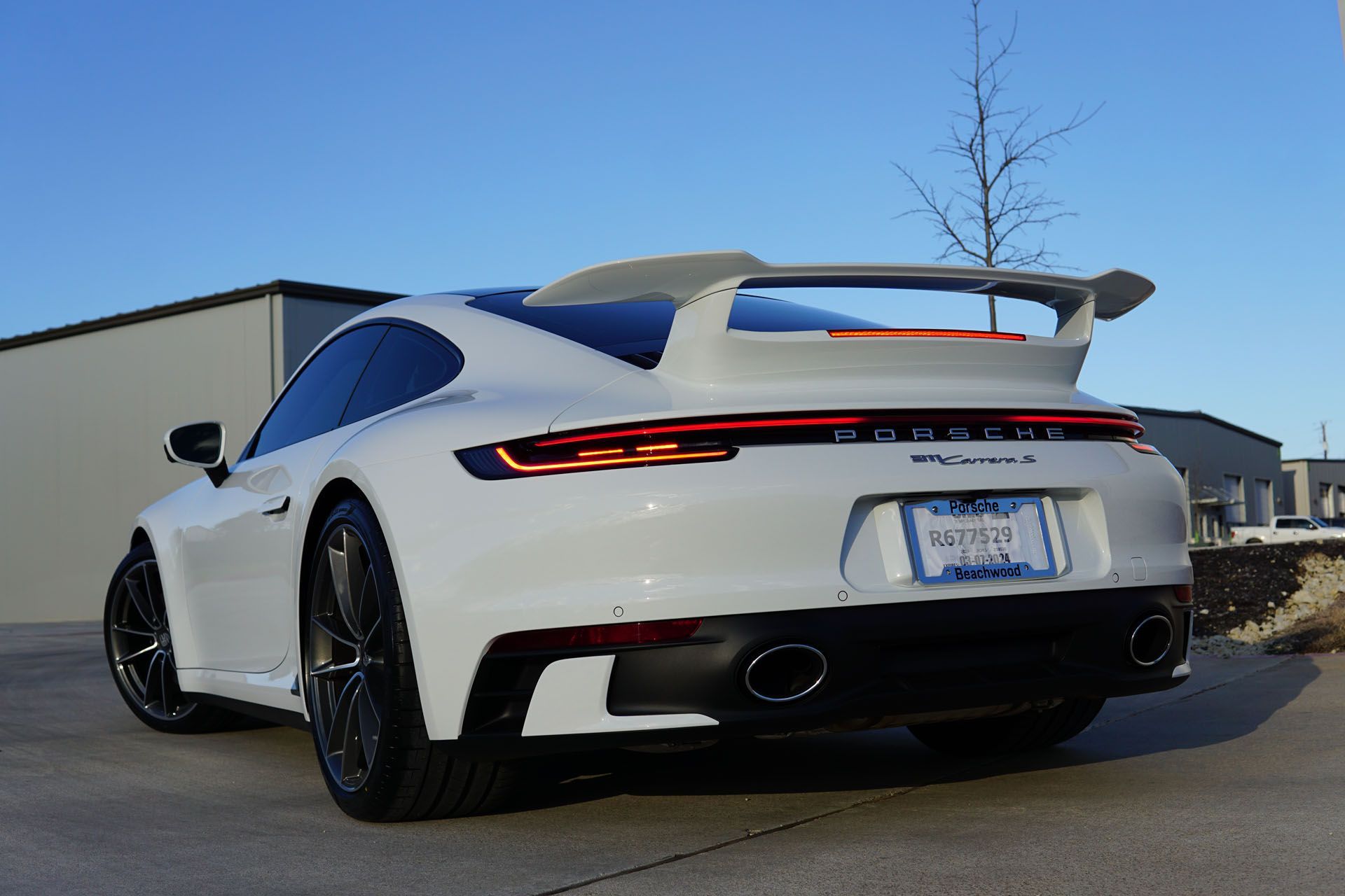 White Porsche 911 Turbo S parked outside on a sunny day. Rear view, black wheels, spoiler, and license plate visible.