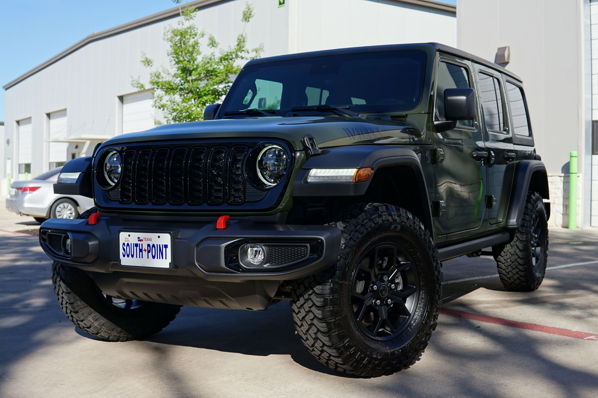 Dark green Jeep Wrangler with black rims and tires parked in front of a light-colored building.