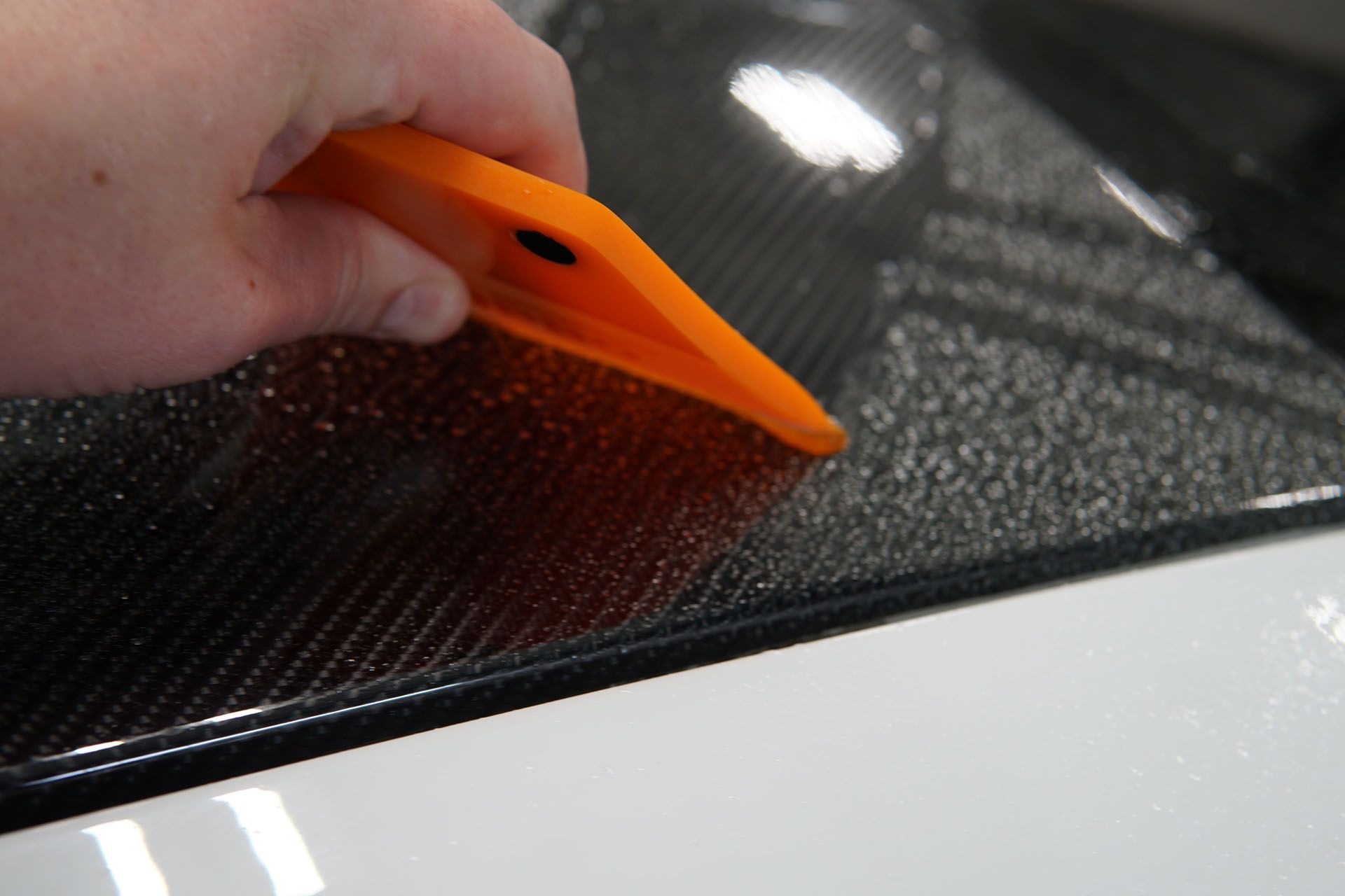 A person is holding an orange squeegee over a car hood.