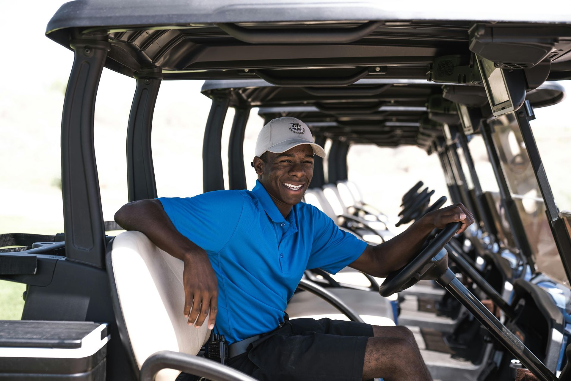 A golf course summer intern in a blue shirt is sitting in a golf cart