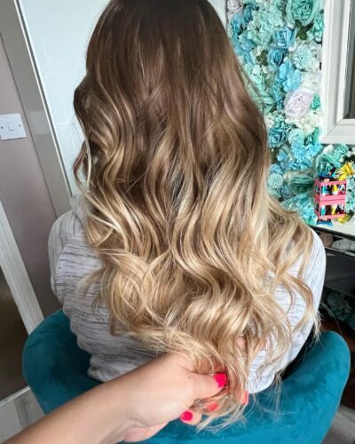 Woman with wavy brown-blonde ombre hair. Hand with pink nails holds the ends. Teal chair and flowered wall background.