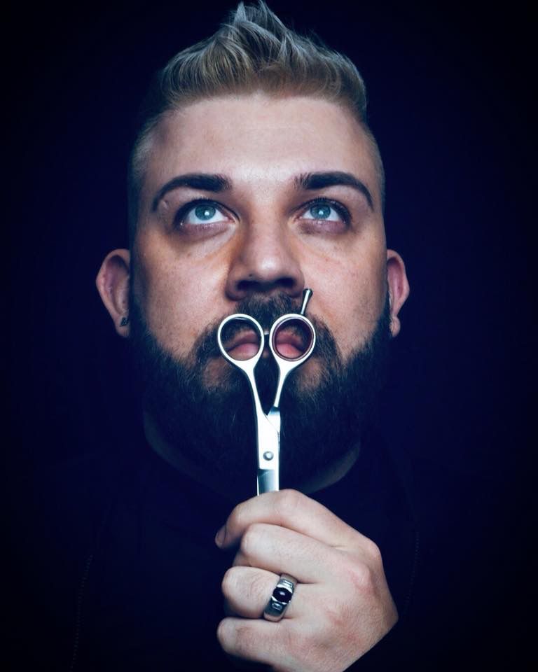 Man with beard, holding scissors up to his nose, looking up, against a dark background.
