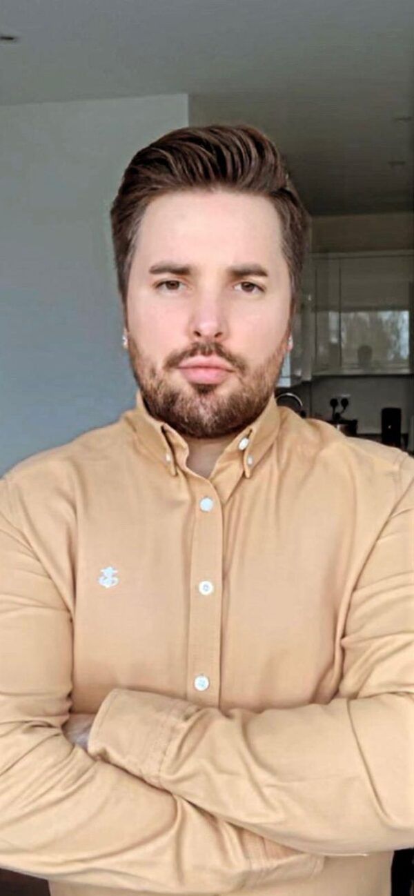 A person with a beard and brown hair, wearing a light orange collared shirt with arms crossed.