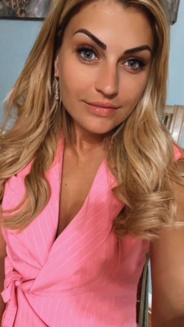 Woman with blonde hair wearing a pink wrap top, looking at the camera.