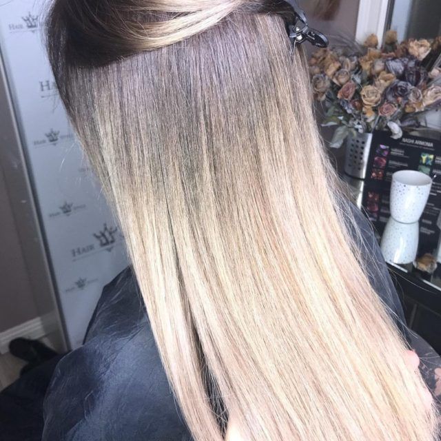 Woman with long blonde hair, hair extensions partially installed, seated in a salon.
