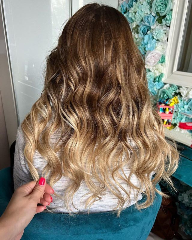 Woman with wavy, blonde-ombre hair, hand holding a strand. Sitting on teal chair, floral wall in the background.
