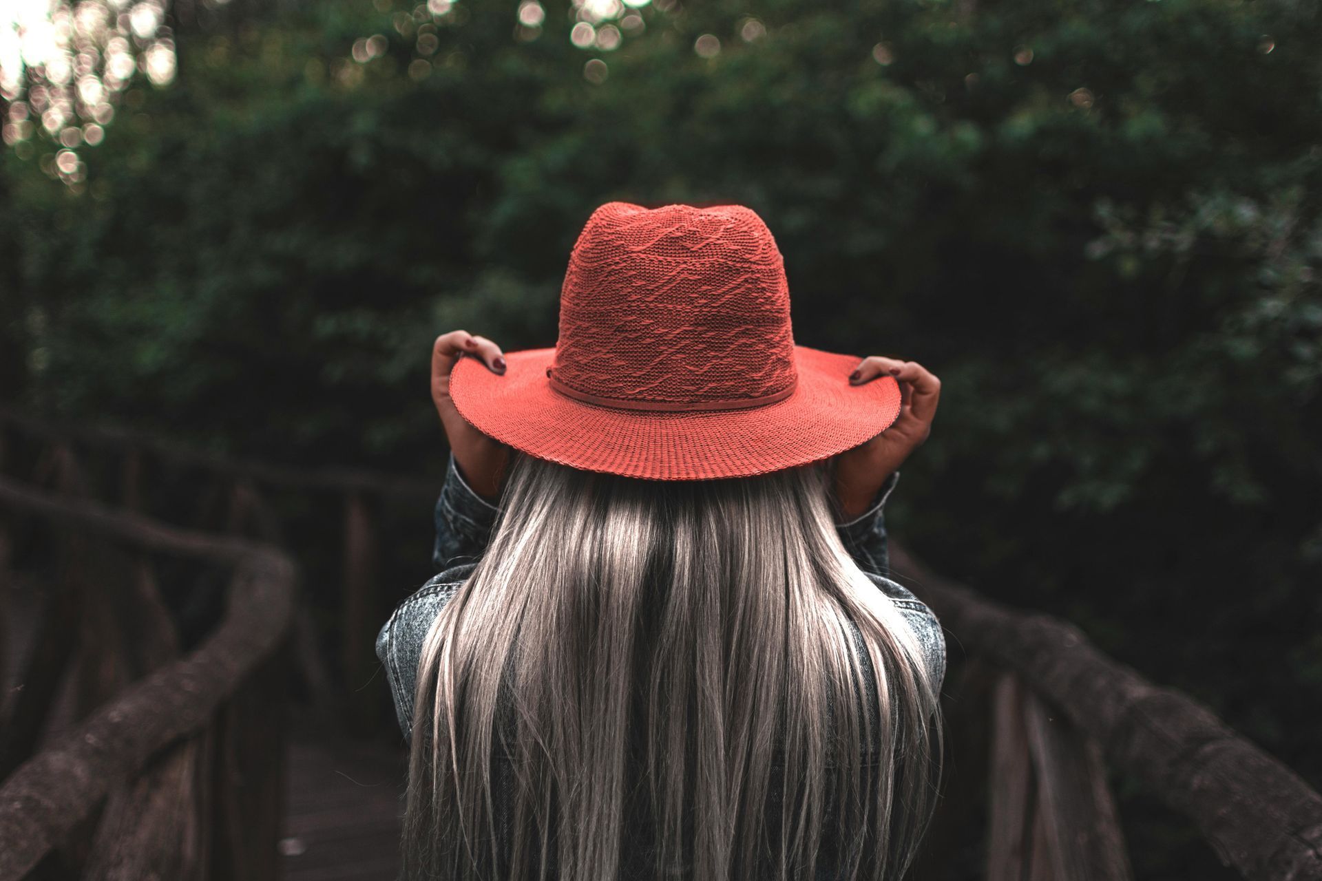 Woman with long gray hair wearing a red hat, standing on a wooden bridge, surrounded by green trees.